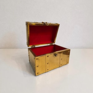 May include: A rectangular brass chest with a hinged lid, featuring a polished gold-tone metallic exterior and a vibrant red interior lining. The box is constructed with visible metal rivets along the edges and includes a central latch mechanism.