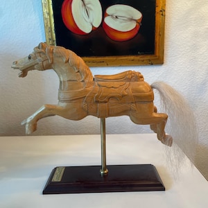 Może przedstawiać: Drewniana rzeźba konia z karuzeli w pozycji skoku, zamontowana na mosiężnym pręcie i ciemnej drewnianej podstawie. Koń jest jasnobrązowy z białym ogonem. Obraz jabłek wisi w tle.