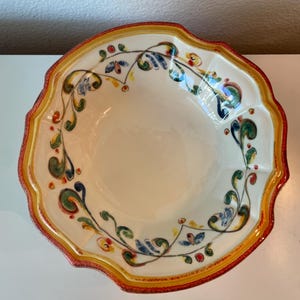 May include: A decorative ceramic bowl with a scalloped edge. The bowl features a cream-colored interior and a vibrant border with red, yellow, and orange accents. Intricate floral and vine patterns in blue, green, and red adorn the rim, creating a colorful design.