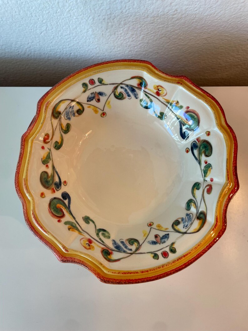May include: A decorative ceramic bowl with a scalloped edge. The bowl features a cream-colored interior and a vibrant border with red, yellow, and orange accents. Intricate floral and vine patterns in blue, green, and red adorn the rim, creating a colorful design.