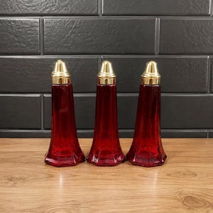 May include: Three matching salt and pepper shakers made of translucent red glass with a faceted, tapered body design. Each shaker is topped with a polished gold-tone metal cap that features a smooth, rounded dome shape and a narrow neck.