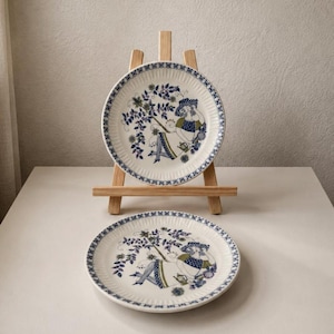 May include: Two round ceramic plates featuring a ribbed edge and a central illustration of a figure resting under a tree. The design uses blue and green tones on a white background. One plate is displayed on a wooden easel, while the other lies flat.
