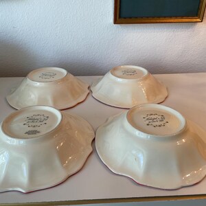 May include: Four off-white ceramic bowls with scalloped edges are arranged on a white surface. The bowls have a red rim and a floral design on the bottom. The bowls are likely for serving food.