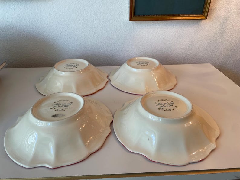 May include: Four off-white ceramic bowls with scalloped edges are arranged on a white surface. The bowls have a red rim and a floral design on the bottom. The bowls are likely for serving food.