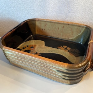 May include: Rectangular ceramic baking dish with a ribbed exterior and integrated handles. The dish features a dark brown interior with a lighter, abstract design. The exterior has a mix of brown, tan, and orange tones.