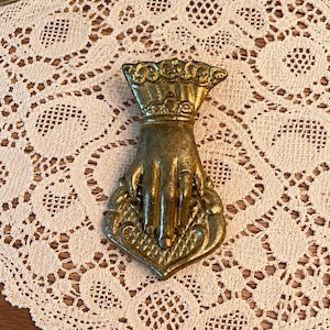 May include: A gold-toned metal clip shaped like a hand with a decorative cuff, holding a heart-shaped base with a textured, cross-hatched pattern. The object features intricate detailing on the wrist and a scalloped edge at the top of the cuff.