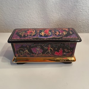 Puede incluir: Una caja decorativa rectangular negra con base dorada. La caja está adornada con ilustraciones coloridas de figuras en varias poses. La tapa y los lados presentan obras de arte detalladas, con un borde dorado que añade un toque de elegancia. La caja está sobre una superficie blanca.