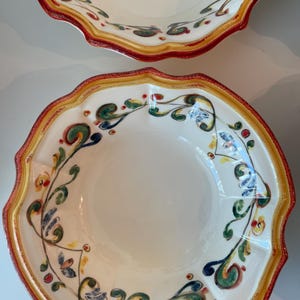 May include: Two off-white ceramic bowls with scalloped edges. The rims are bordered with a red, yellow, and green design. The bowls feature a floral pattern in green, blue, and red. The bowls are likely for serving food.