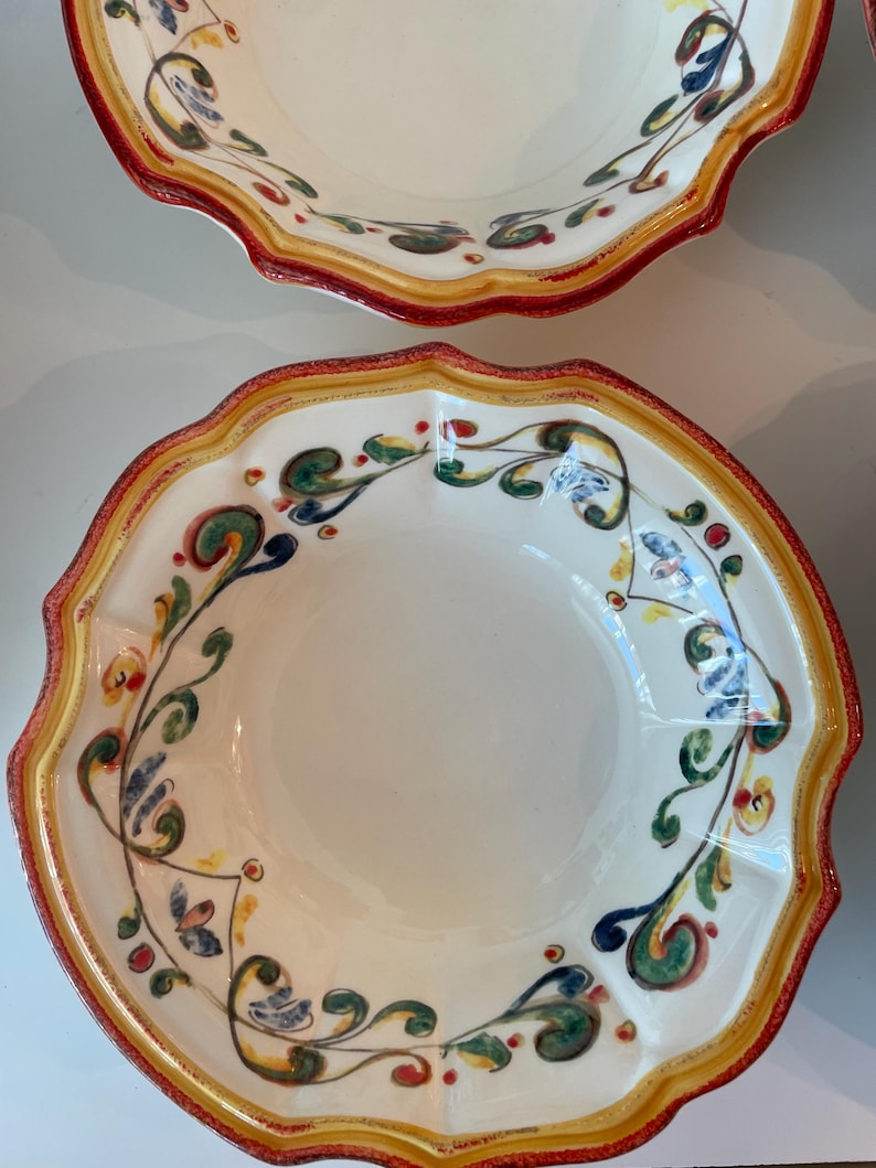 May include: Two off-white ceramic bowls with scalloped edges. The rims are bordered with a red, yellow, and green design. The bowls feature a floral pattern in green, blue, and red. The bowls are likely for serving food.