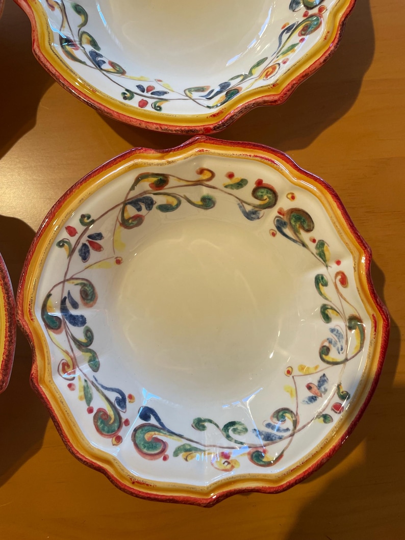 May include: A set of white ceramic bowls with a scalloped edge. The rims are decorated with a red and yellow border, and the interior features a colorful floral design in green, blue, and yellow.