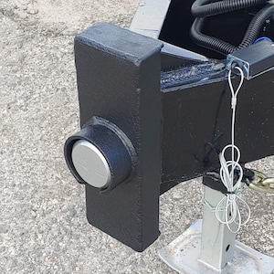 May include: Close-up of a black metal trailer component with a silver cap and a silver jack stand. The component has a textured surface and is mounted on a concrete surface. A white wire is attached to the component.