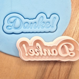 Könnte beinhalten: Zwei Ausstecher, einer blau und einer rosa, beide mit dem Wort "Danke!" in einer kursivschrift.