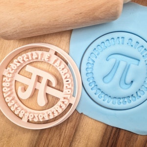 Op de afbeelding: Een roze plastic koekjesvormpje in de vorm van het wiskundige symbool pi, met de cijfers van pi rond de rand. Het koekjesvormpje ligt op een blauw oppervlak met een deegroller op de achtergrond.