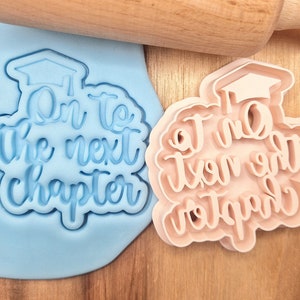 Peut inclure: Deux emportes-pièces en plastique bleu et rose avec le texte "On to the next chapter" en écriture cursive. Les emportes-pièces sont en forme de chapeau de graduation.