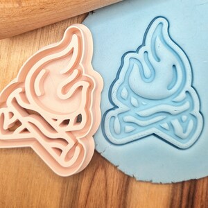 May include: A pair of cookie cutters, one pink and one blue, shaped like a campfire with flames. The cutters are made of plastic and are designed for baking and decorating cookies.