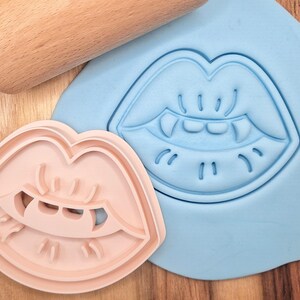 May include: Two pink and blue cookie cutters shaped like a pair of lips with fangs. The cookie cutters are on a light blue surface.