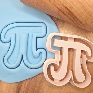 Può includere: Due stampini per biscotti a forma del simbolo matematico pi greco. Lo stampino a sinistra è blu ed è premuto nell'argilla da modellare blu. Lo stampino a destra è rosa e si trova su una superficie di legno.