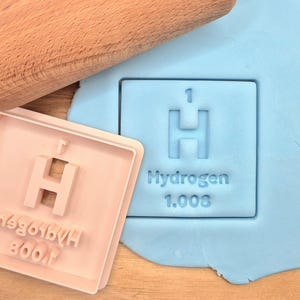May include: A pink plastic cookie cutter shaped like a periodic table element, with the symbol "H" for Hydrogen, the atomic number "1", and the atomic weight "1.008".
