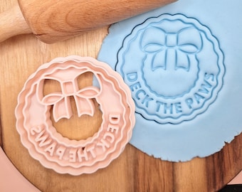 Puede incluir: Dos cortadores de galletas de color rosa y azul con forma de corona y un lazo en el centro. Los cortadores tienen el texto "Deck the Paws" alrededor del borde.