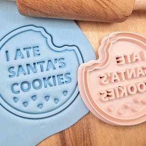 May include: A blue and pink cookie cutter with the text "I ate Santa's cookies" on it. The cutter is designed to make a cookie in the shape of a bitten cookie.