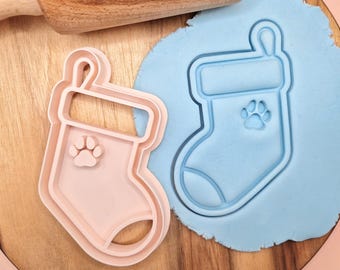 Puede incluir: Dos cortadores de galletas de plástico rosa en forma de calcetín de Navidad con un diseño de huella de pata. Los cortadores están sobre una superficie de madera con plastilina azul.
