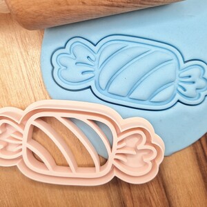 Op de afbeelding: Een roze plastic koekjesvormpje in de vorm van een snoepstok op een blauw oppervlak met een houten deegroller op de achtergrond.