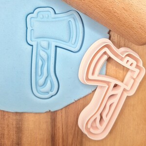 May include: Two plastic cookie cutters in the shape of an axe. One cutter is blue and the other is pink.