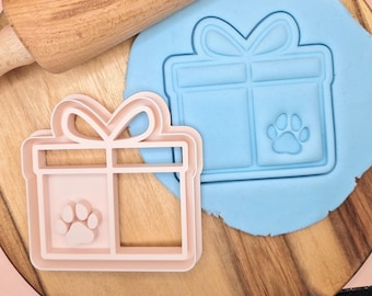 Puede incluir: Dos cortadores de galletas de plástico rosa en forma de cajas de regalo con un diseño de huella de pata. Los cortadores están sobre una superficie de madera con un rodillo azul y una pieza de arcilla para modelar azul.