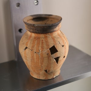 May include: A small, broken, orange and brown clay pot with a striped pattern. The pot has several holes in it.
