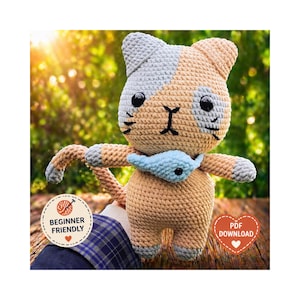 May include: Crocheted cat doll in shades of tan, gray, and blue. The cat has black eyes and a blue fish-shaped accessory around its neck. The doll is held in a person's hand. The image includes a "Beginner Friendly" and "PDF Download" logo.