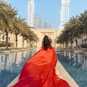 Può includere: Una persona con un lungo abito rosso si allontana dalla fotocamera, con il tessuto che si muove dietro di lei. L'ambientazione è uno spazio esterno lussuoso con palme, una piscina ed edifici moderni, tra cui un grattacielo sullo sfondo.