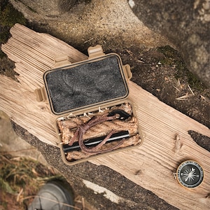 Könnte beinhalten: Ein beigefarbenes Survival-Kit mit einem schwarzen Feuerstarter, Zunder und einer Lederschnur. Das Kit ist geöffnet und zeigt den Inhalt. Ein Kompass befindet sich neben dem Kit. Die Gegenstände liegen auf einem Stück verwittertem Holz.