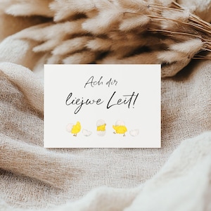 Könnte beinhalten: Eine cremefarbene Karte mit dem Text "Ach dir, Liewe Leit!" in schwarzer Schrift, mit Aquarellillustrationen von gelben Küken, die aus Eiern schlüpfen. Die Karte befindet sich vor einem neutral getönten, strukturierten Hintergrund mit getrockneten Pflanzen.