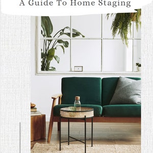 May include: A green velvet sofa with a gray pillow sits in a living room with a large window. A small round table with a woven top and black metal legs sits in front of the sofa. The table has a clear glass top and a glass bottle on top.