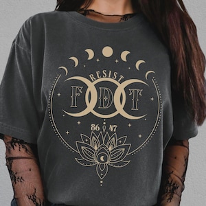 May include: Dark gray t-shirt with a gold graphic design featuring the word "RESIST" above the letters "FDT" inside a circle with crescent moons and a lotus flower. The shirt has a relaxed fit.