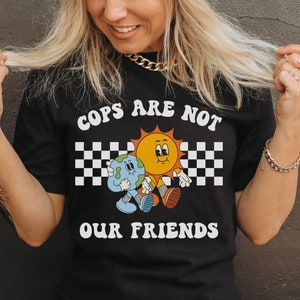 May include: A black t-shirt with white text that reads 'Cops Are Not Our Friends'. The text is in a retro font and is surrounded by a checkered pattern. A cartoon sun and earth are holding hands and walking in the checkered pattern.