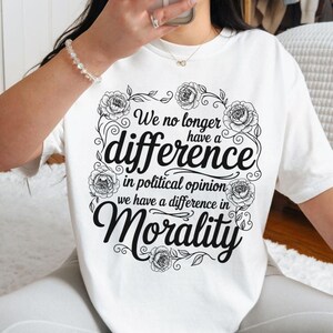 May include: White t-shirt with a black floral design surrounding the words "We no longer have a difference in political opinion, we have a difference in Morality." The shirt has short sleeves and a crew neck.