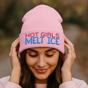 May include: A light pink beanie with the text "HOT GIRLS MELT ICE" in blue and red. The beanie is worn by a person with long brown hair and a pink sweatshirt. The background is blurred, with a path and trees visible.