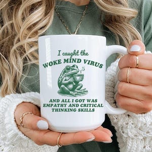 May include: White ceramic mug with a green frog illustration and the text "I caught the woke mind virus and all I got was empathy and critical thinking skills".