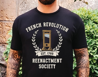 May include: A black t-shirt with a white graphic design featuring a guillotine, laurel wreath, and the text "French Revolution Est. 1789 Reenactment Society".