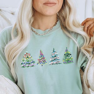 Könnte beinhalten: Hellgrünes Sweatshirt mit vier gestickten Weihnachtsbäumen. Jeder Baum ist mit bunten Ornamenten geschmückt. Die Bäume sind in Rosa-, Blau-, Grün- und Gelbtönen gehalten. Das Sweatshirt hat lange Ärmel.