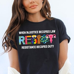 Pode incluir: Camiseta preta com o texto "Quando a injustiça se torna lei, Resista. A resistência se torna dever." A palavra "Resista" é composta por símbolos coloridos, incluindo um punho, um útero, um arco-íris e um globo.