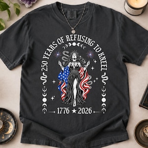 May include: A charcoal gray t-shirt featuring a graphic of a figure draped in an American flag, surrounded by snakes, stars, and moon phases. The text reads 250 YEARS OF REFUSING TO KNEEL and 1776 2026, with a central star separating the two dates.