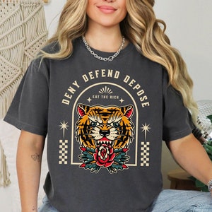 May include: A dark gray t-shirt with a graphic of a tiger's face in a vintage tattoo style. The text "Deny Defend Depose Eat The Rich" is printed around the tiger's face.