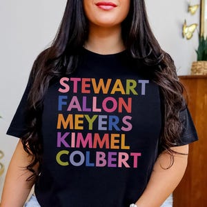 May include: Black t-shirt with the names STEWART, FALLON, MEYERS, KIMMEL, and COLBERT in colorful block letters. The shirt is a crew neck and is being worn.