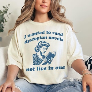 May include: A woman wearing a white t-shirt with a blue graphic of a woman reading a book. The text on the shirt reads "I wanted to read dystopian novels not live in one."