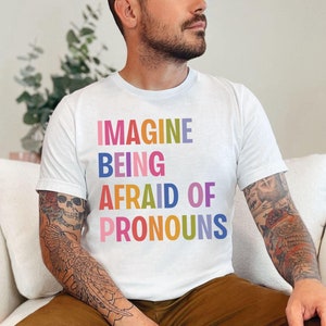 May include: A white t-shirt with the text "IMAGINE BEING AFRAID OF PRONOUNS" printed in rainbow colors. The text is stacked vertically, with each word in a different color.