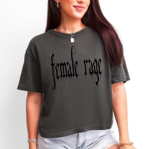 May include: Dark grey t-shirt with the words "female rage" in a gothic font. The shirt has short sleeves and a relaxed fit. The text is in black.