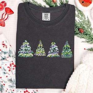 May include: Dark gray crew neck t-shirt featuring four Christmas tree designs with colorful ornaments. The trees are embroidered with white, purple, yellow, and black details. The shirt is surrounded by holiday decorations.