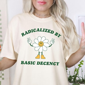 Könnte beinhalten: Ein weißes T-Shirt mit einer grünen Grafik einer lächelnden Gänseblümchen mit Armen und Beinen. Die Gänseblümchen trägt gelbe Schuhe. Der Text "Radicalized by Basic Decency" ist in Grün unter der Gänseblümchen gedruckt.
